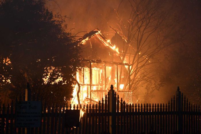 Fire engulfs the front of a house. The front fence is silhouetted by the flames.